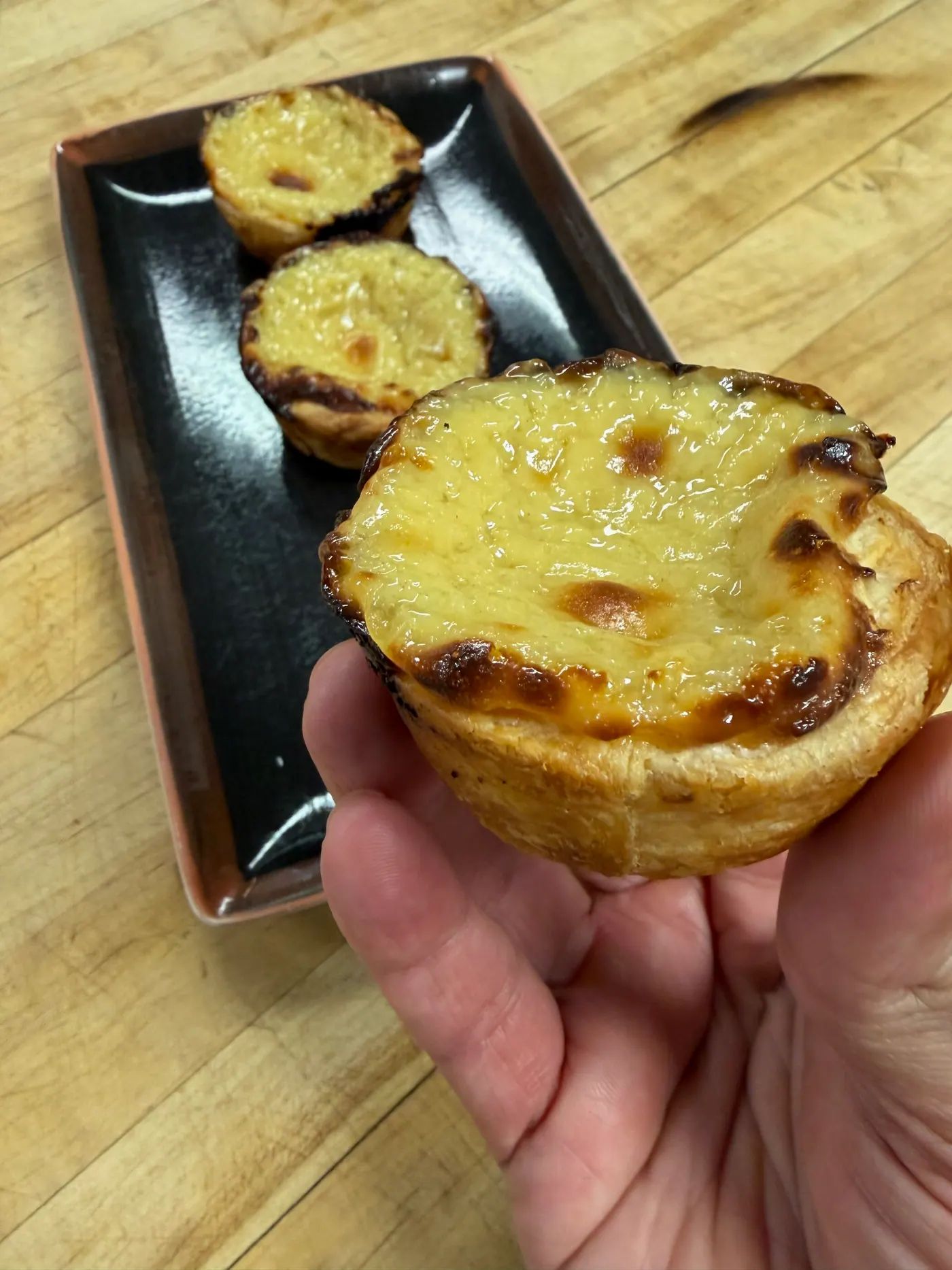 A hand holding a golden custard tart over a tray of more